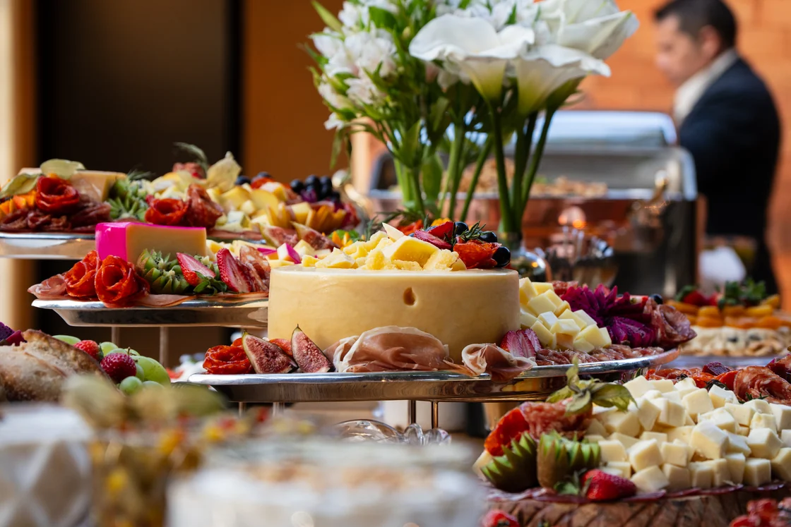 Mesa de buffet com queijos e frutas, com uma pessoa desfocada ao fundo